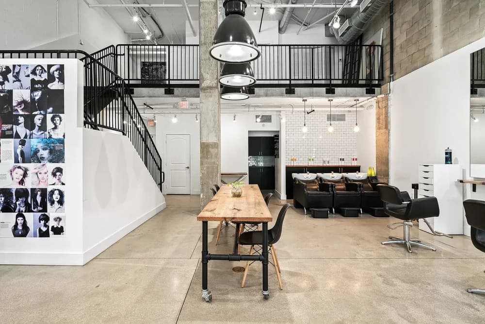 The main floor of Scotch Bonnet Hair Studio, a photo-collage wall, a communal wood table and the black steel staircase in a double-height downtown West Palm Beach loft.