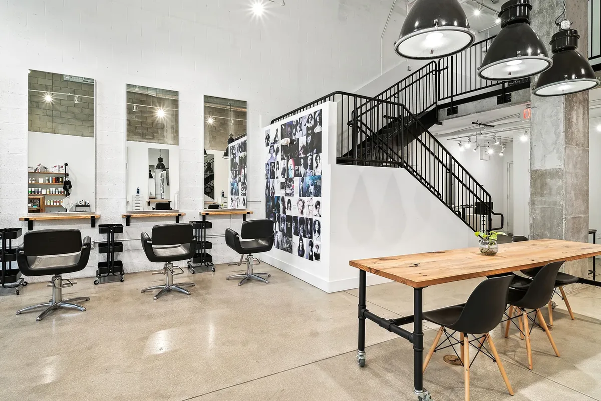 The communal table under industrial pendant lamps and the loft staircase inside the downtown West Palm Beach hair studio.